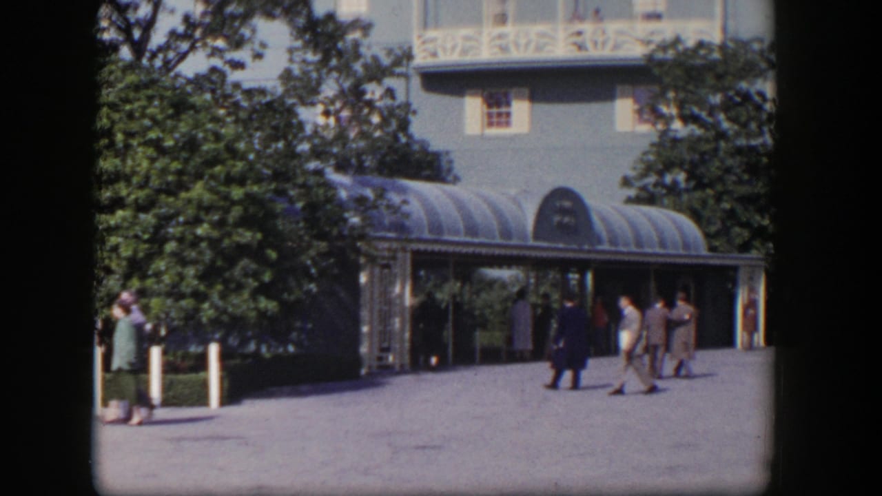1954 Las Vegas home-movie frame showing visitors near a covered resort entrance, blue hotel walls, white trim, trees, and bright sun.