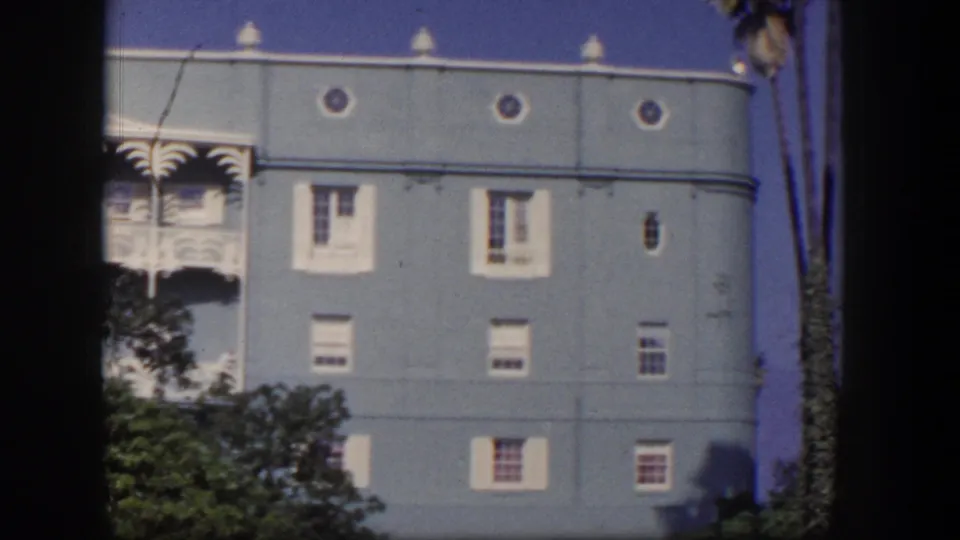 1954 Las Vegas resort exterior with blue walls, decorative white balconies, small windows, foliage, and palm trees along the edge.