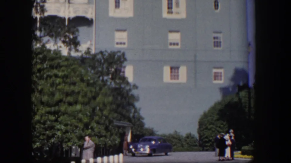 1954 Las Vegas resort driveway with visitors, a dark 1950s-era car, white bollards, trees, palms, and the blue hotel facade behind them.
