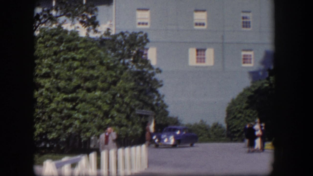 1954 Las Vegas home-movie frame showing a light-blue hotel, trees, pedestrians, and a deep blue car near the entrance road.