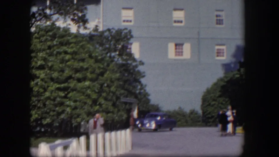 1954 Las Vegas home-movie frame showing a light-blue hotel, trees, pedestrians, and a deep blue car near the entrance road.