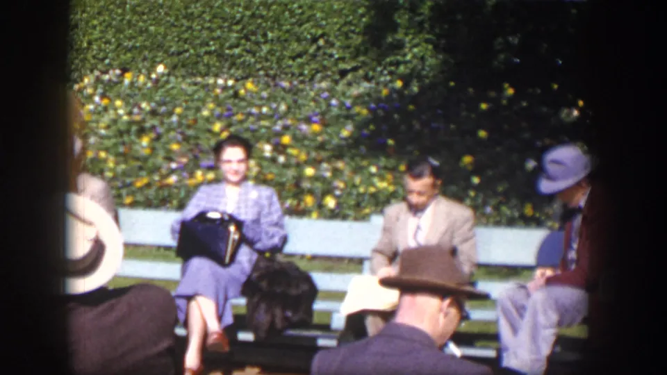 Well-dressed adults sit on benches in a Las Vegas garden setting, reading and talking amid greenery and flowers.