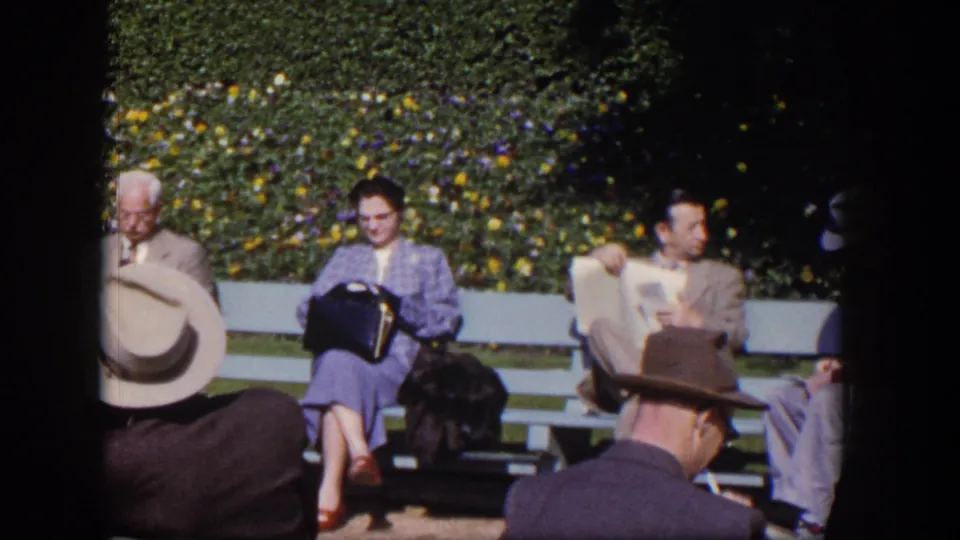 Adults in 1950s attire sit on garden benches in Las Vegas, with hats, newspapers, and dense flowering shrubs behind them.