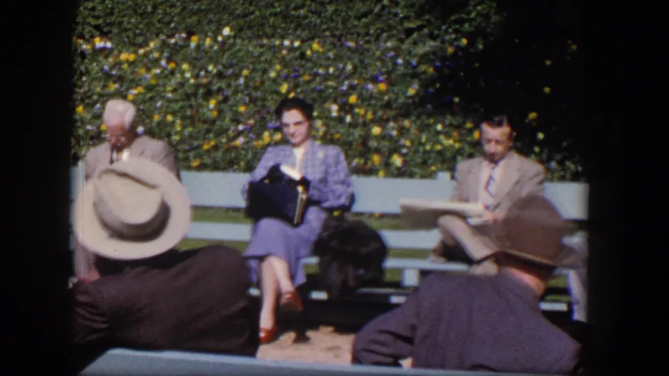 Adults in mid-century attire sit on Las Vegas park benches in 1954, framed by foreground hats and a hedge with small flowers.