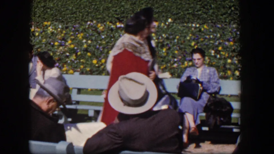 1954 Las Vegas park footage shows a passerby crossing in front of seated adults reading and resting near a flowering hedge.