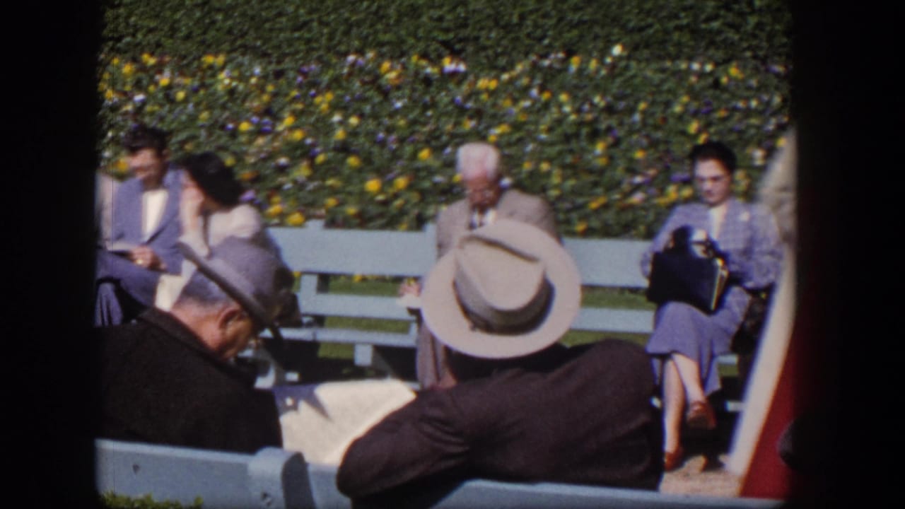 Adults in suits, dresses, and hats sit on light blue benches beside a flowering hedge.