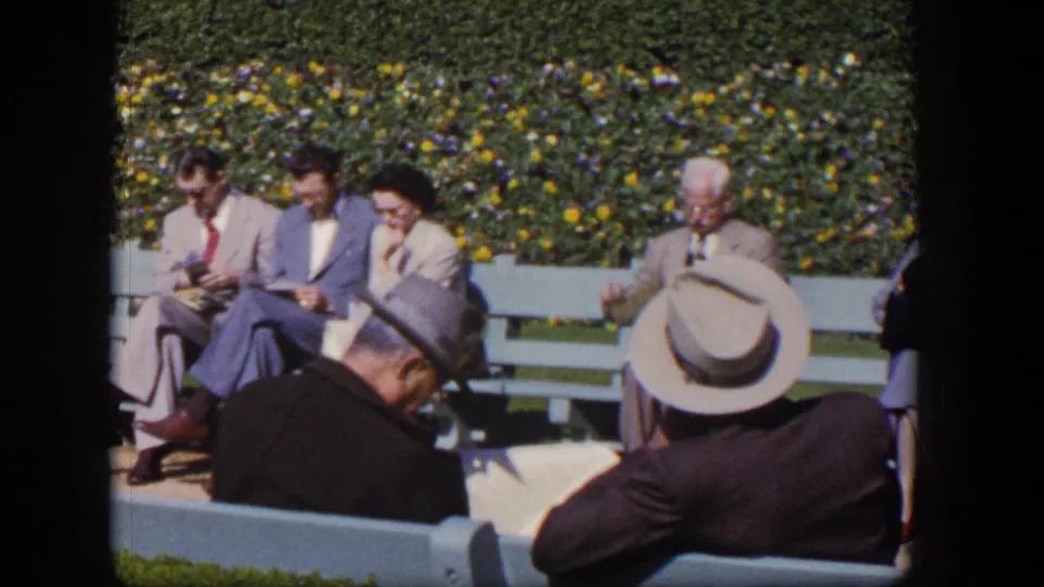 Adults in mid-century dress sit on light blue benches in a sunny Las Vegas garden setting, with faces visible.