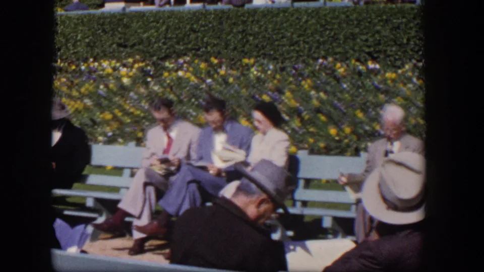 Home-movie view of adults seated outdoors in 1954 Las Vegas, with suits, hats, benches, and a flowering hedge visible.