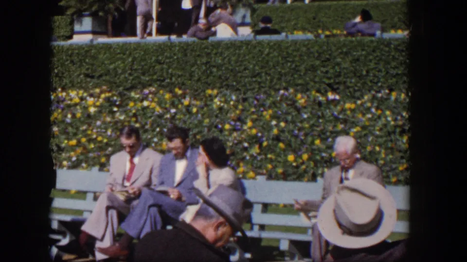 1954 Las Vegas 8mm frame with adults in social clothes seated on benches, partly obscured by nearby hat-wearing figures.