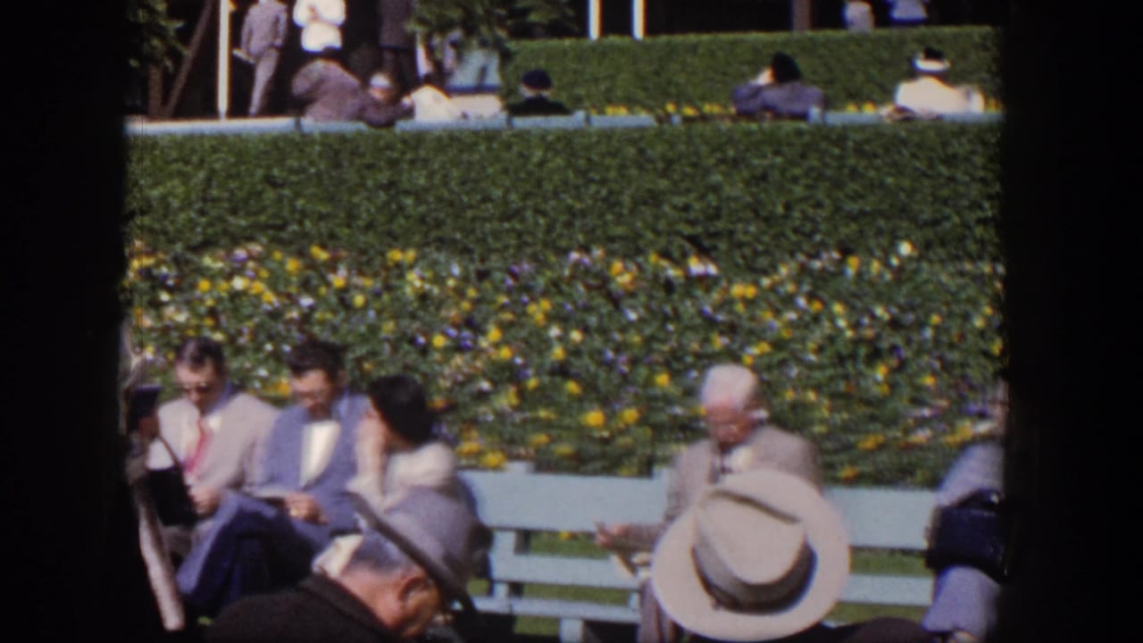 1954 Las Vegas home-movie frame showing adults in suits, hats, and dresses around benches beside a planted hedge.