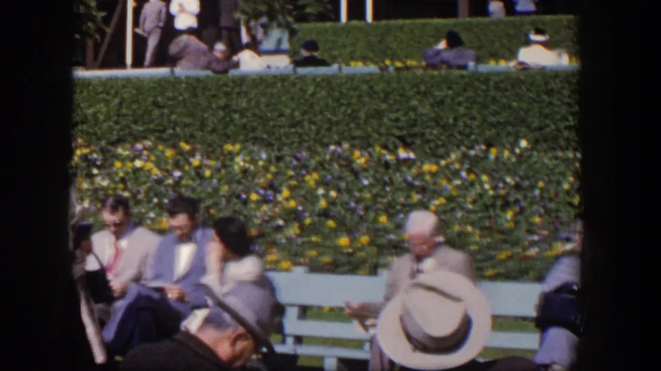 1954 Las Vegas home-movie frame showing adults in suits, hats, and dresses around benches beside a planted hedge.