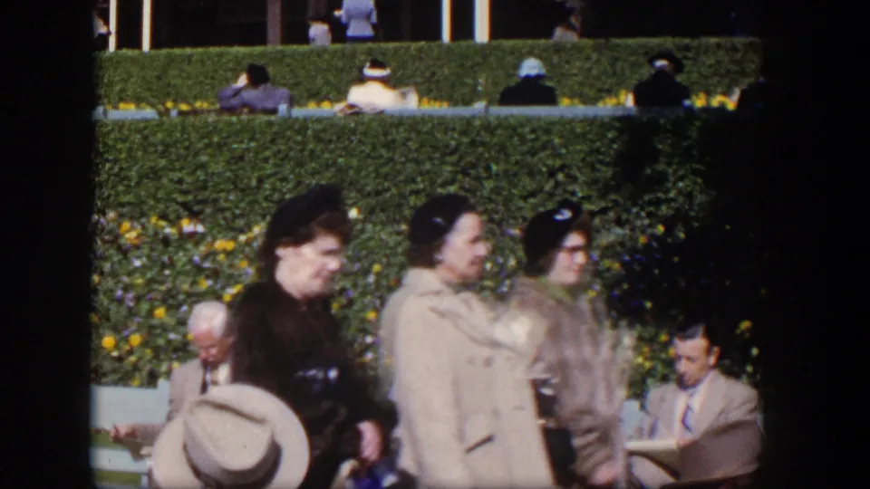 1954 Las Vegas home-movie frame showing women walking past seated adults, with hats, coats, benches, and flowers visible.