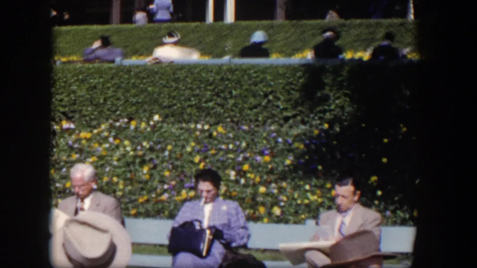 1954 Las Vegas frame showing seated adults on benches, including people reading, with flowers and hedges in the background.
