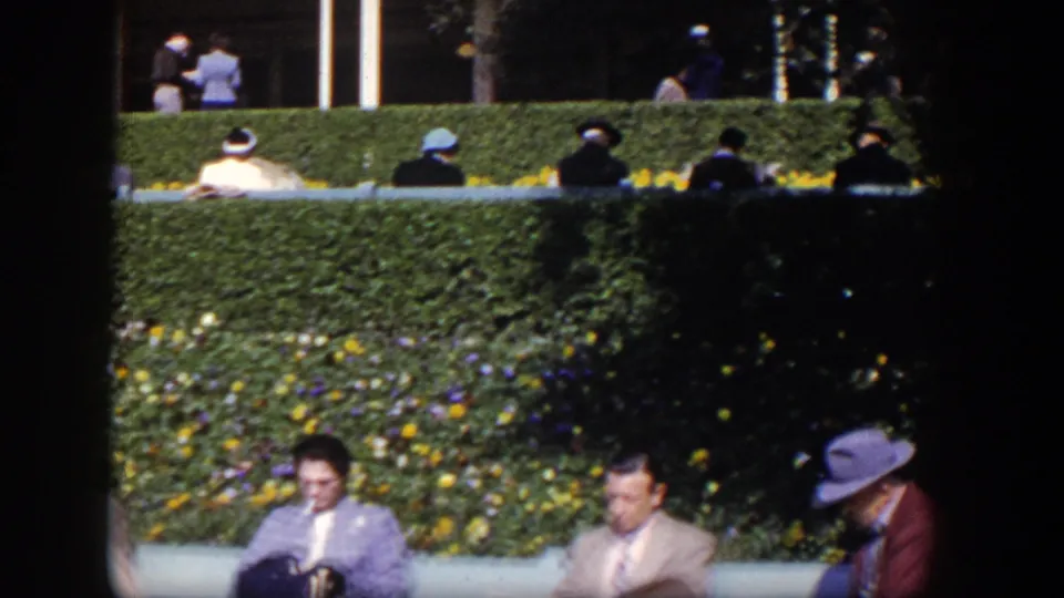 1954 Las Vegas home-movie frame with suited adults and women in hats seated by manicured hedges and flowers.