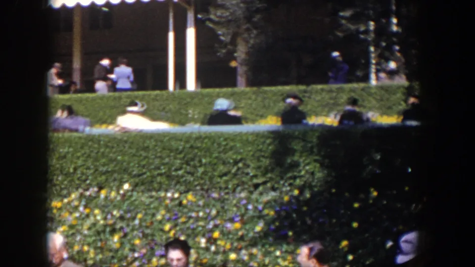 Las Vegas, 1954 frame showing adults seated behind clipped hedges and flowerbeds under a shaded awning edge.