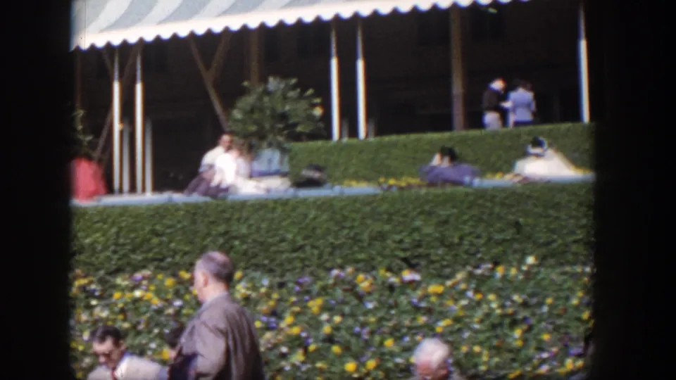 Late-frame 1954 Las Vegas view of seated adults behind a hedge, with flowers and a suited man in the foreground.