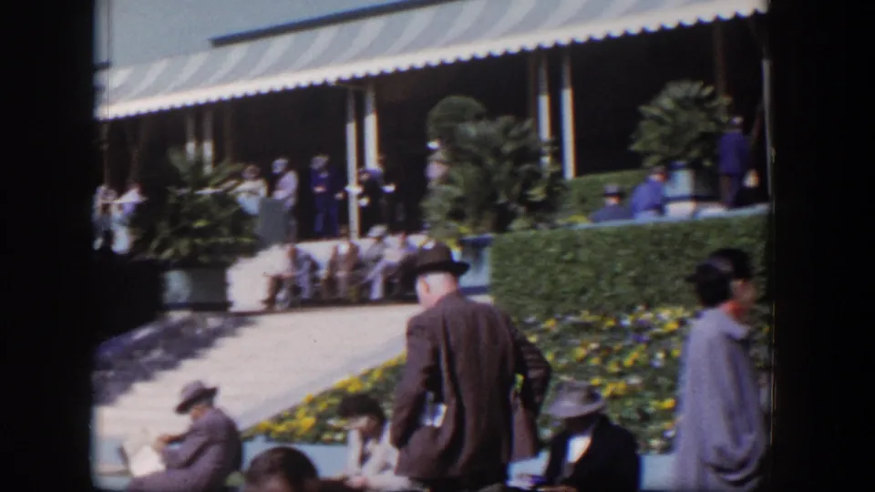 A wider 1954 Las Vegas racetrack view shows spectators moving and sitting around hedges, flowers, and shaded seating.