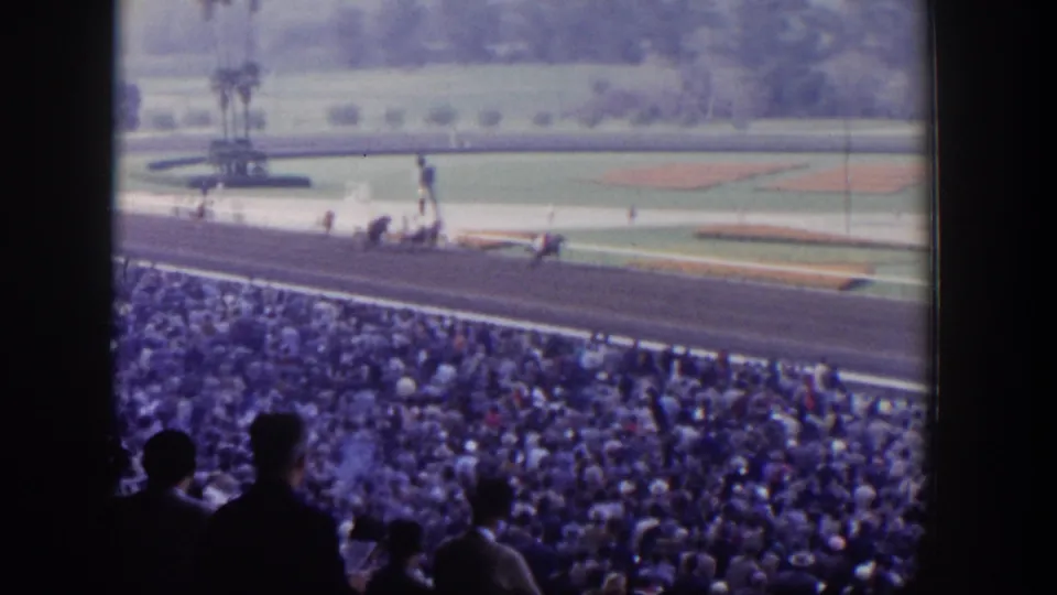 1950s racetrack action in Las Vegas, showing horses on the near stretch, spectators in shadow, and landscaped infield beyond.