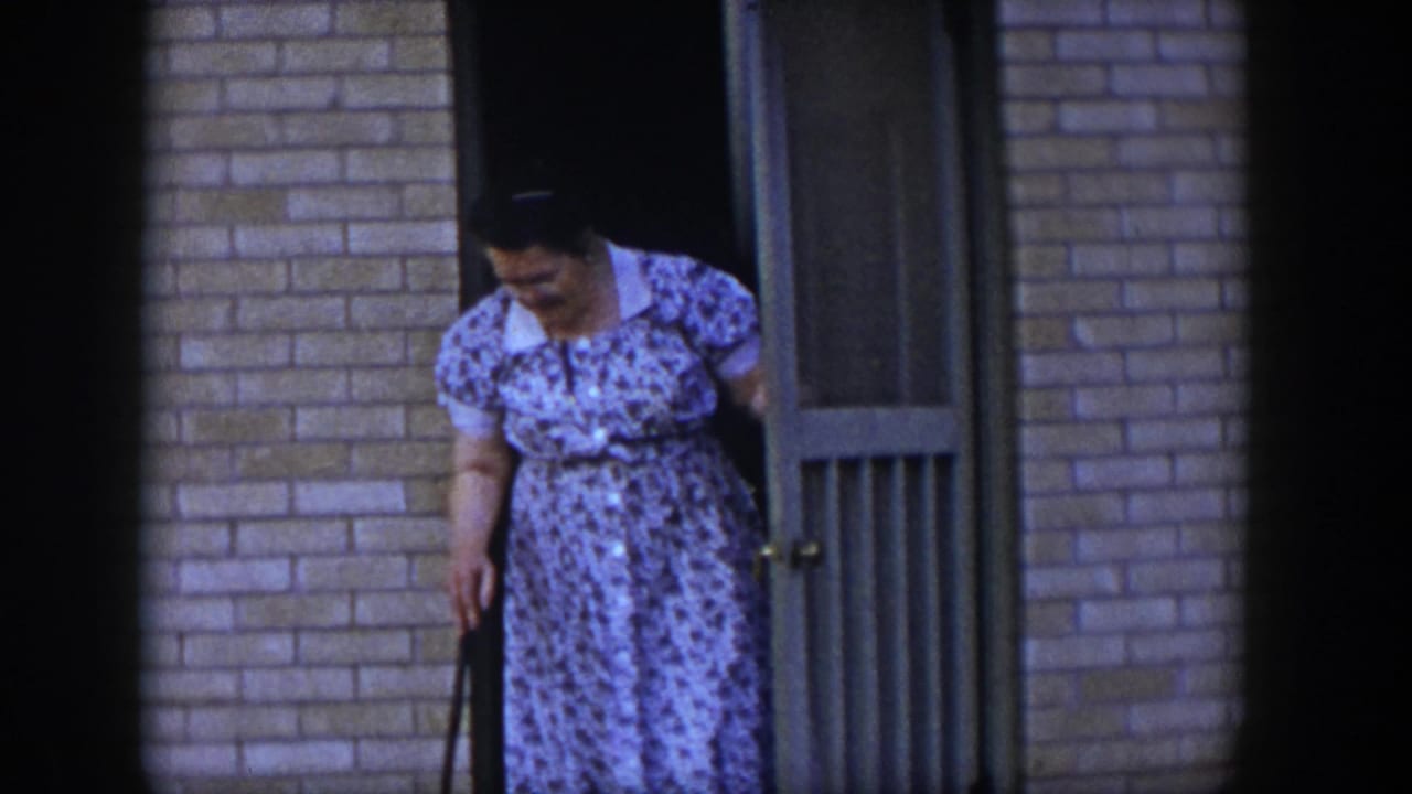 In 1954 Las Vegas home-movie footage, a woman in a blue floral dress steps from a tan screen door beside a brick wall.