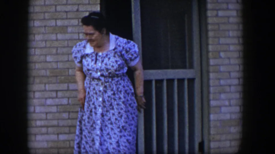 The woman in a blue floral dress stands just outside the tan screen door of a brick home in 1954 Las Vegas footage.