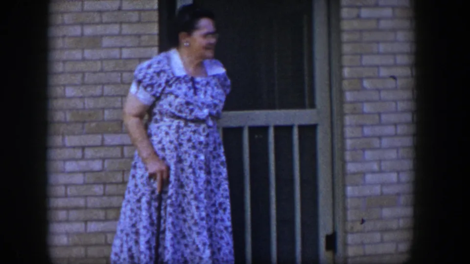 A woman holding a cane moves slowly near the brick exterior and tan screen door in this 1954 Las Vegas home movie.