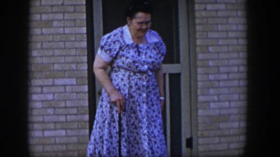 Near the tan screen door, the woman in a blue floral dress leans forward while holding a cane in 1954 Las Vegas footage.