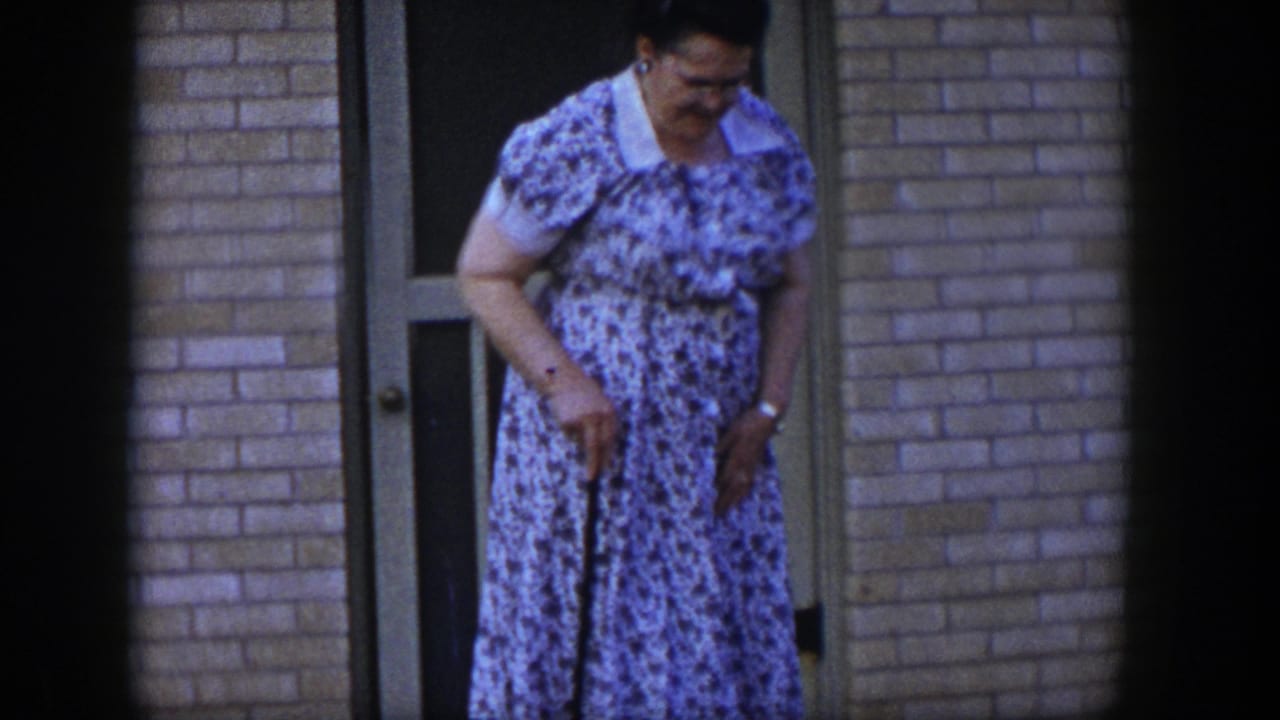 Older woman in a floral dress stands near a doorway with a walking stick.