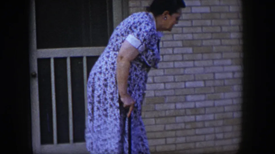 In the Las Vegas 1954 home-movie frame, the woman leans on a walking stick while moving along the brick exterior.