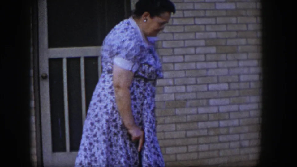 The older woman, wearing a floral dress and glasses, moves carefully past a brick doorway in the 1954 Las Vegas footage.