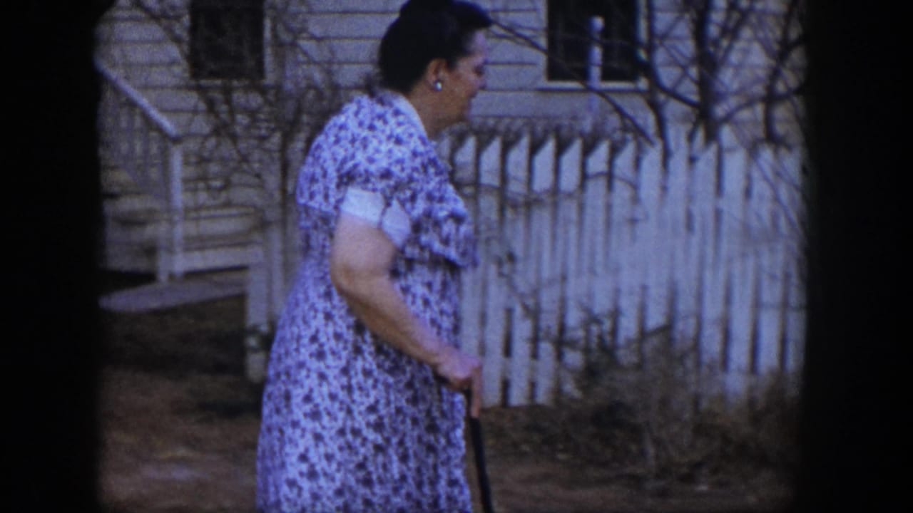 In 1954 Las Vegas home-movie footage, a woman in a floral dress walks slowly with a cane near a white picket fence and house exterior.
