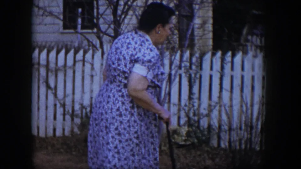 The woman continues through the Las Vegas residential yard, using a cane as bare shrubs and a white fence fill the background.