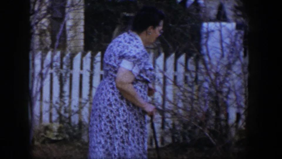 The woman looks toward the ground while moving with her cane in the residential yard, with fence slats and bare branches behind her.