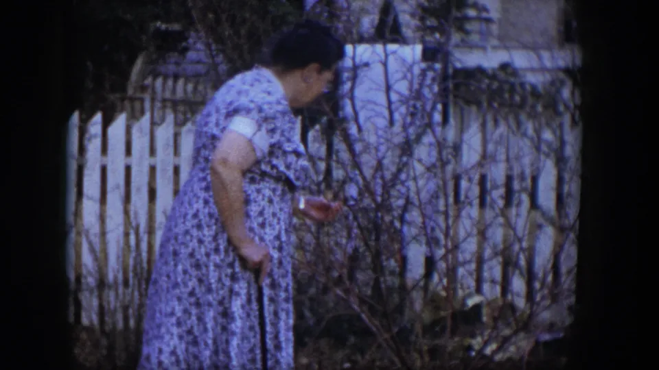 Near the end of the clip, the woman pauses or slows with her cane among bare yard branches, with a white fence and house exterior behind her.
