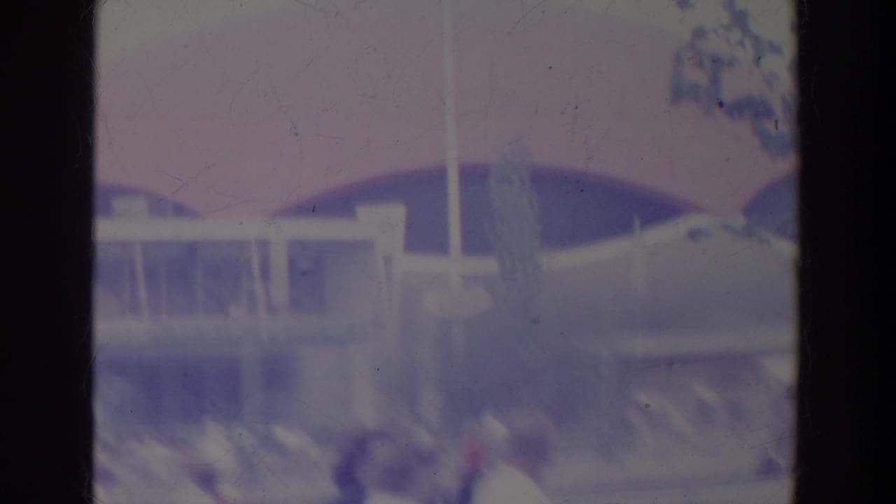 1964 World's Fair home-movie frame showing fountain spray, modern structures, visitors, and part of the Unisphere in soft purple-blue color.