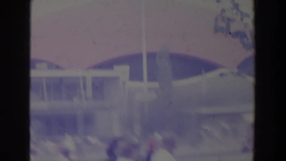 1964 World's Fair home-movie frame showing fountain spray, modern structures, visitors, and part of the Unisphere in soft purple-blue color.