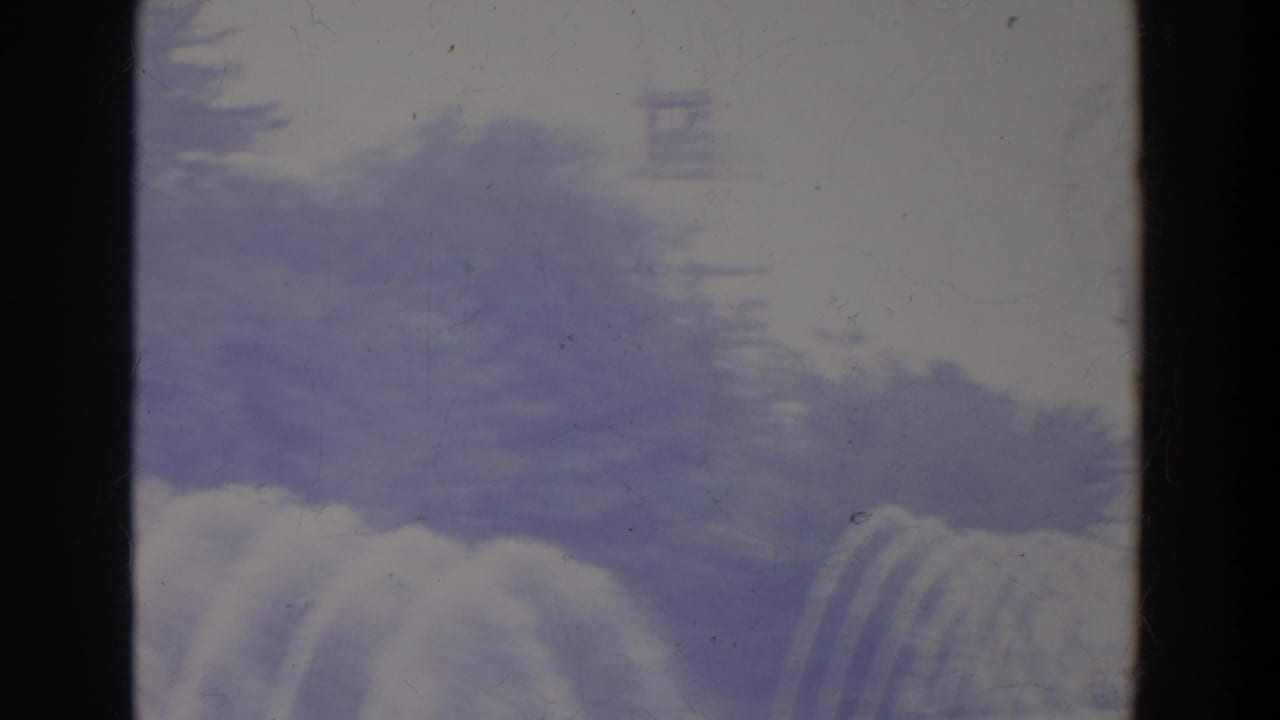 1964 8mm home-movie frame with hazy sky, soft pale forms, and dark vertical framing, with heavy grain and scratches visible.