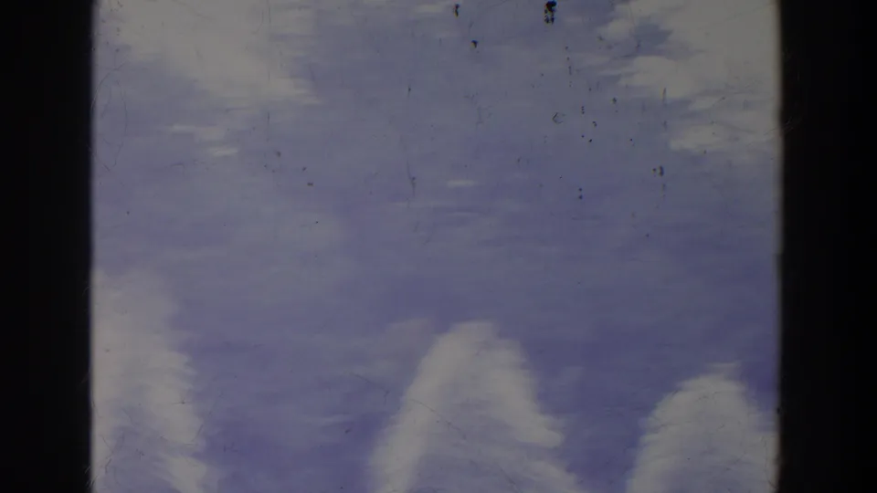 Hazy 8mm frame showing blue-purple sky, soft white cloud or spray forms, and dark vertical divisions, with dust and scratches across the film.