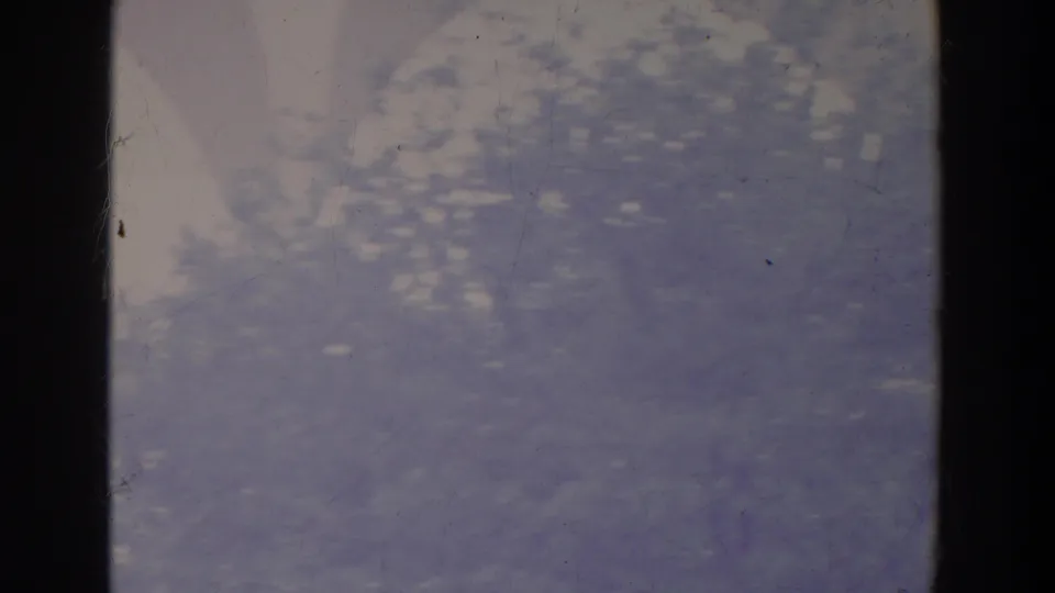 Soft 1964 8mm frame with blue-gray water or mist, a pale diagonal shape, dark vertical framing, and visible dust, scratches, and color fading.