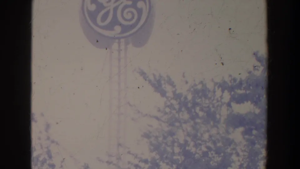 Faded 8mm view of a GE logo sign raised above trees, with pale sky and partial fair structures below.
