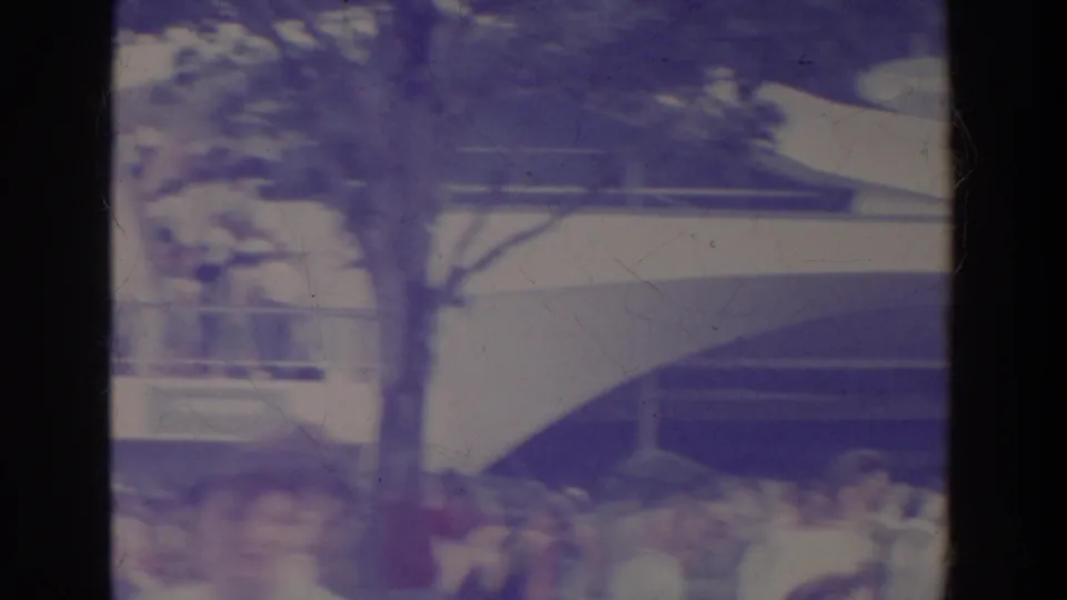 Faded 8mm frame showing a crowd gathered beneath a white curved fair building, with railings, trees, and partial signage area above.