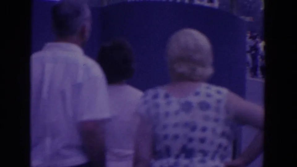 Visitors, including a man in a white shirt and a woman in a blue polka-dot dress, walk past a blue fairground wall.