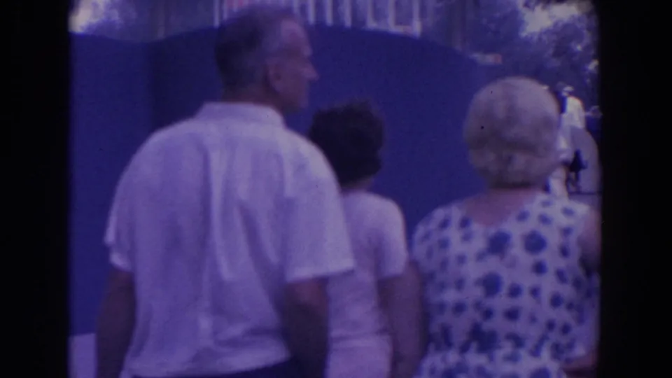 Home-movie frame of adults walking away from the camera beside a blue fairground wall, with a patterned dress prominent in the foreground.