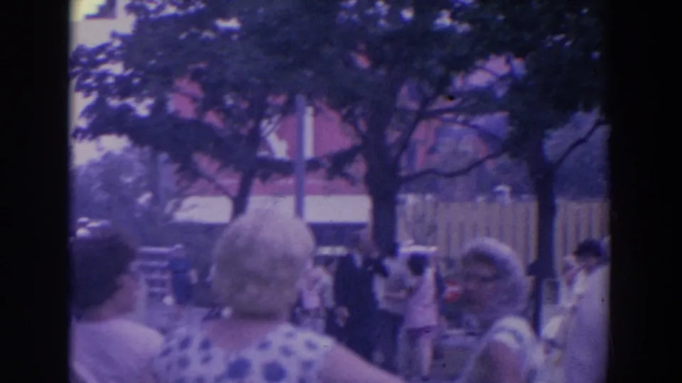 Visitors gather along a tree-lined fairground walkway with fencing and a building visible beyond the crowd.