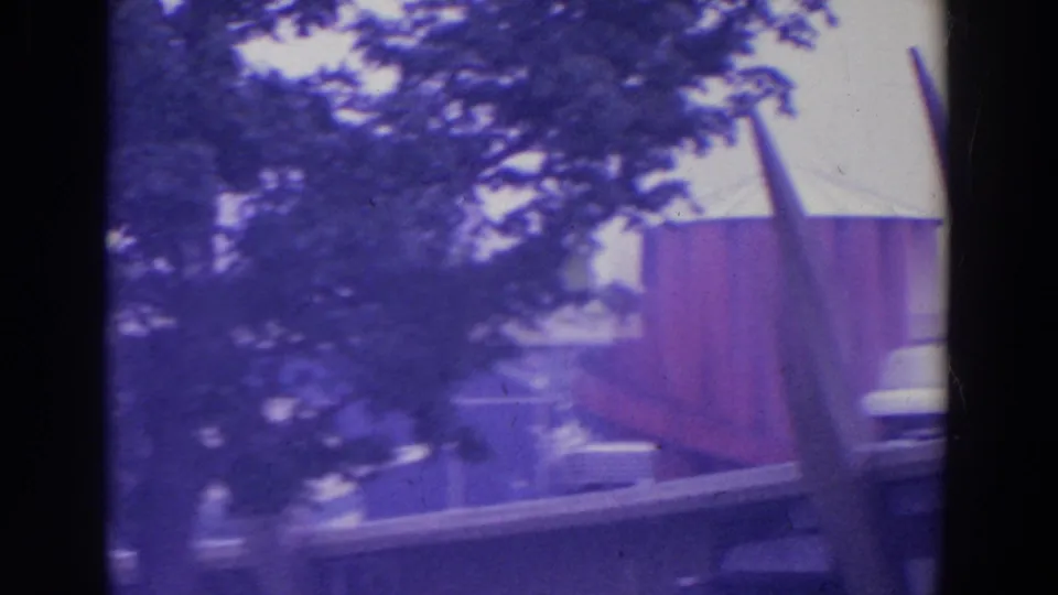 Handheld 8mm view of a pointed fairground form, red-paneled structure, trees, and railings at the 1964 New York World's Fair.