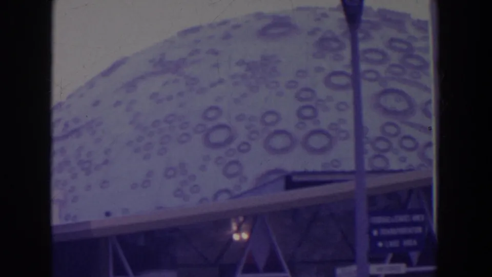 Visitor-shot 8mm frame of a patterned dome-like fair structure, vertical pole, railings, and small signs at the 1964 New York World's Fair.