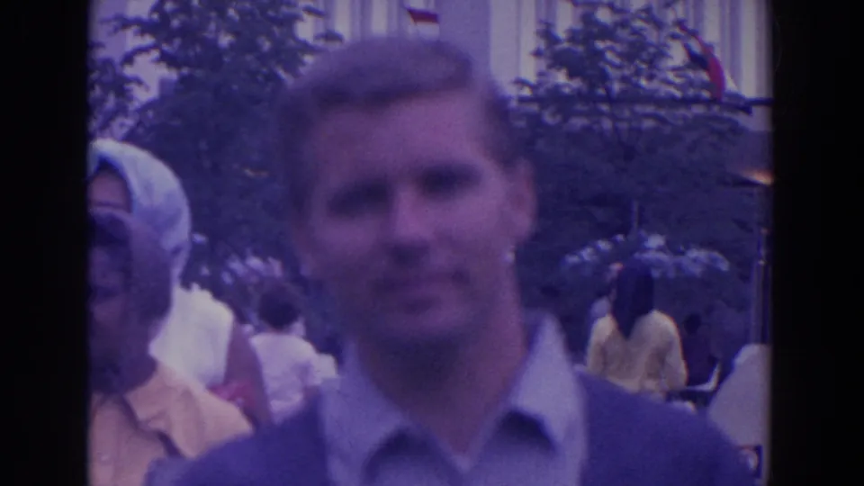 A man in a collared shirt and sweater stands close to the camera, with fairgoers and trees in the background.