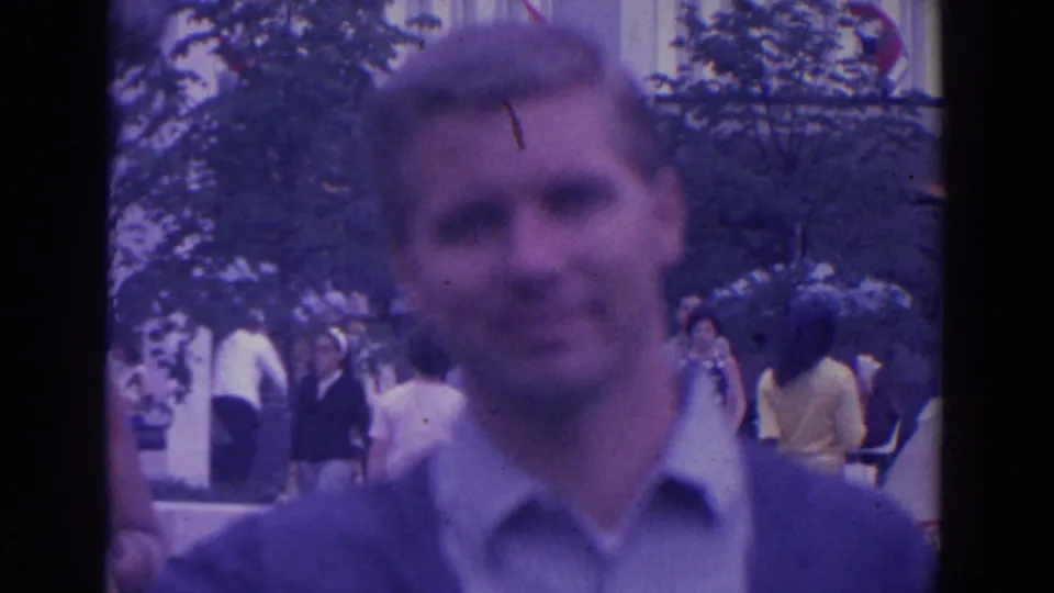 The foreground man is heavily soft-focused while several visitors move through a tree-lined fairground walkway behind him.