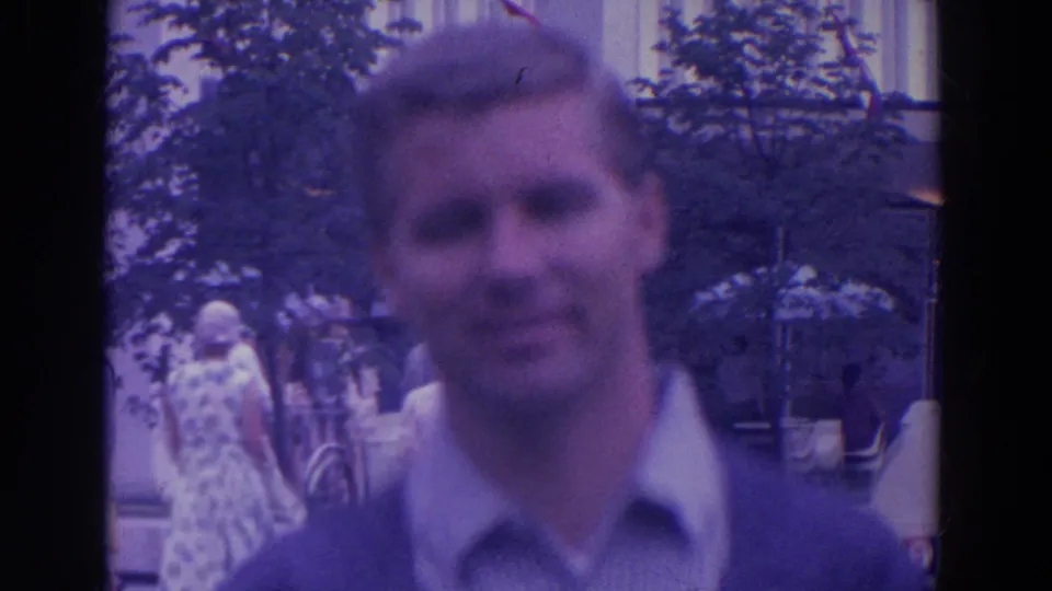 A soft-focus man faces the camera as visitors, trees, and bicycles fill the public fairground space behind him.