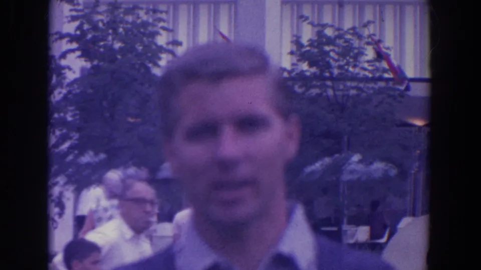 Soft 8mm frame of a man near the lens with adult visitors, trees, and fairground architecture in the background.