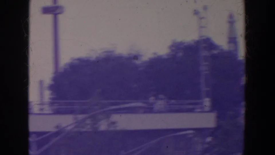 Handheld 8mm view across a fairground walkway with railings, trees, vertical towers, and indistinct visitors.